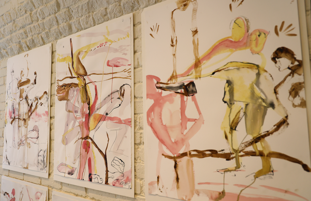 Three paintings with pink, green, and brown streaks and figures against a white-bricked wall.